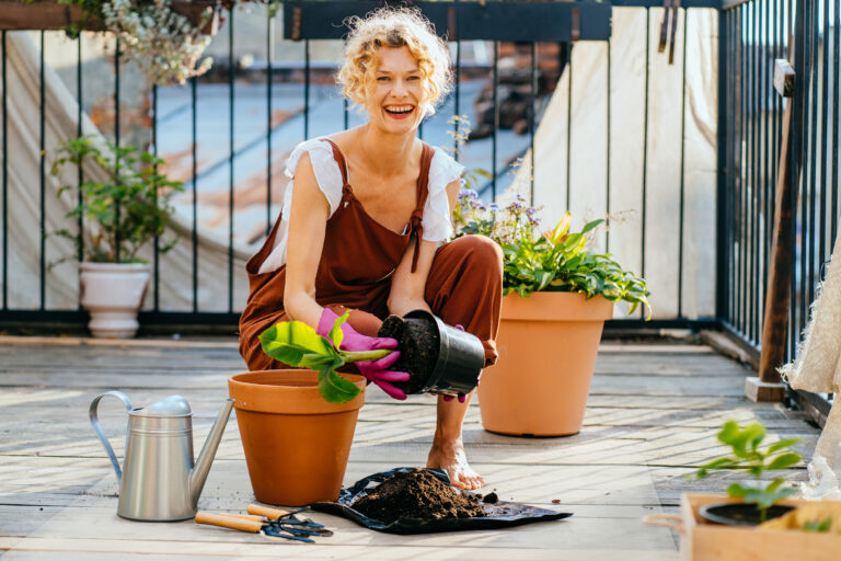 Beautiful amazing blond romantic woman among flowers at her balcony during planting. People, gardening, planting and profession concept. Gardener planting banana tree to flower pot on terrace at home.